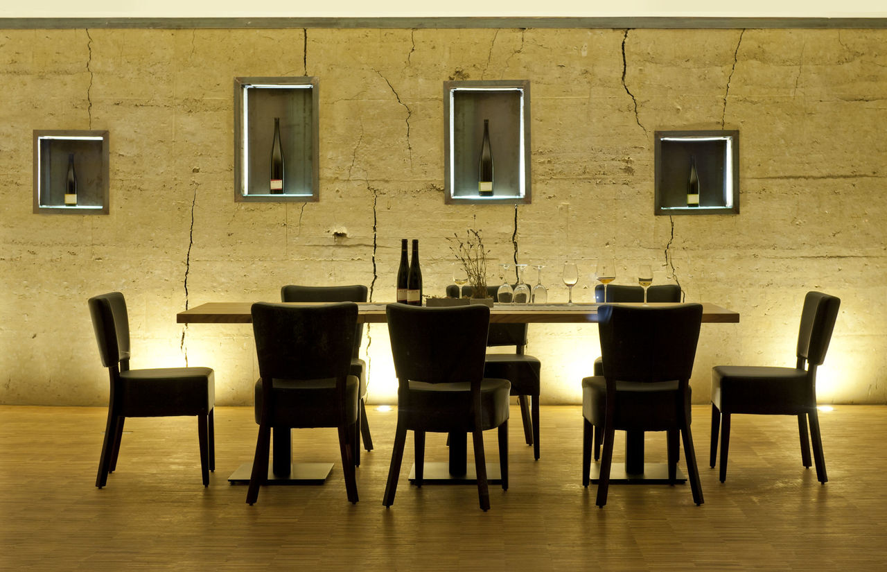 Foto: In die beleuchtete Stampflehmwand sind unterschiedlich große Nischen eingelassen in denen der Wein präsentiert wird, davor befindet sich ein dunkler Holztisch mit passenden Stühlen