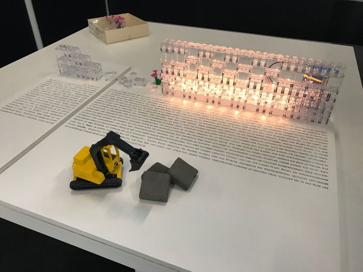 Blick auf ein Modell aus beleuchteten, durchsichtigen Legosteinen. Im Vordergrund ein Spielzeugbagger, im Hintergrund Steinchen mit Kunstblumen.