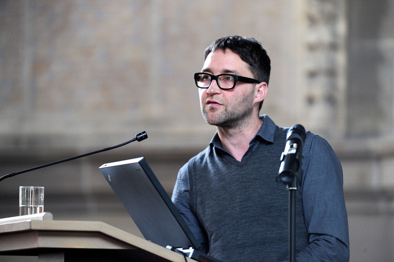 Mann mit Brille und schwarzem Haar spricht in ein Mikrofon vor einer grauen Steinfassade.