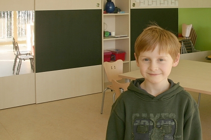 Junge im Grundschulalter steht fröhlich lächenld im Klassenraum.