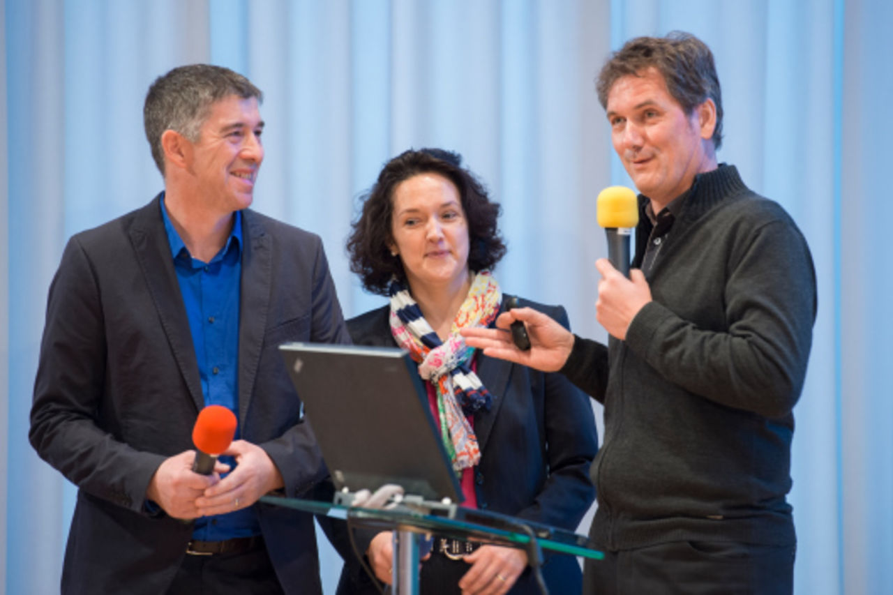Zwei Männer mit Mikrofonen und eine Frau stehen hinter einem Rednerpult