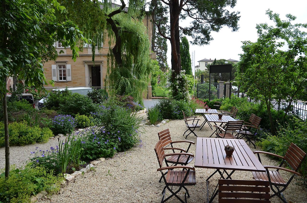 Foto: Blick auf die Außensitzplätze, die sich in einem mit Sträuchern und Bäumen bewachsenen Garten befinden, der Garten ist mit Kieselsteinen angelegt.