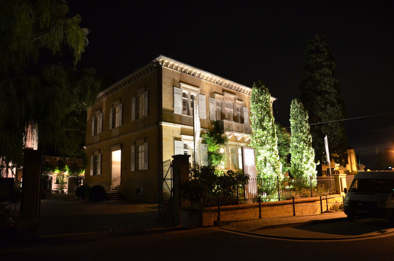 Foto: Beleuchtetes Gebäude bei Nacht. Vor dem Gebäude stehen zwei angestrahlte Zypressen.