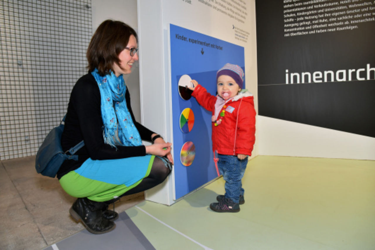 Ein etwa einjähriges Kind steht an einer Wand der Dauerausstellung und spielt am Mitmachangebot "Experimentieren mit Farbe", daneben kniet die Mutter und lächelt. das Kind an