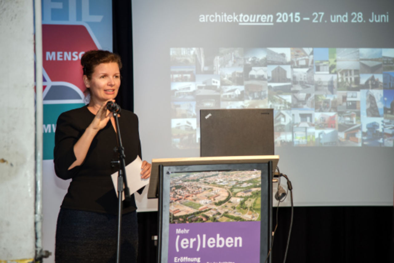 Eine Frau steht am Rednerpult und umfasst das Mikrofan am Mikroständer. Im Hintergrund ein Projektionsbild mit dem Titel "achitektouren 2015" mit einem Mosaikbild aus vielen Architekturprojekten.