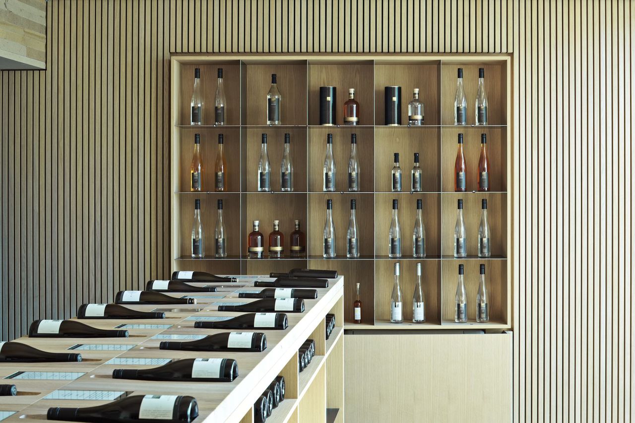 Foto: Rückwand des Verkaufsraumes mit hellen Holzlamellen verkleidet und mit integrierter, rechteckiger Wandvitrine. Davor befindet sich ein weiteres Regal zu Weinpräsentation.