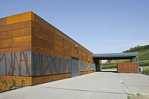 Foto: Blick auf den Betriebshof, mit Rostbrauner Fassade die im unteren Bereich durch einem Lochblech mit Rebenoptik verziert ist, im Hintergrund liegen die Weinberge.