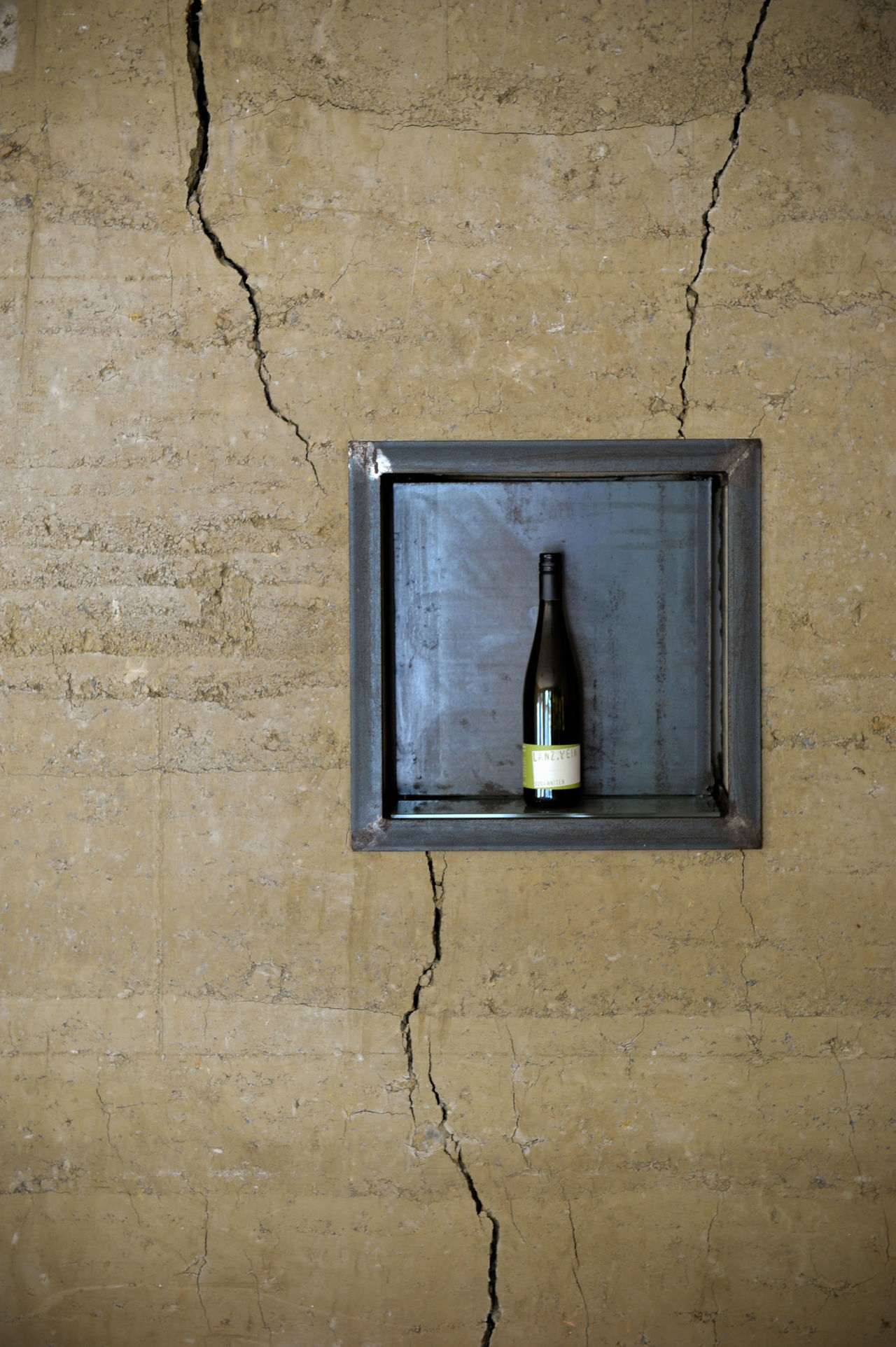 Foto: Aus Lehm gestampfte Rückwand die Trockenrisse als Gestaltungsmaterial aufweist. In die Wand ist ein quadratisches Weinregal aus Eisen  integriert.