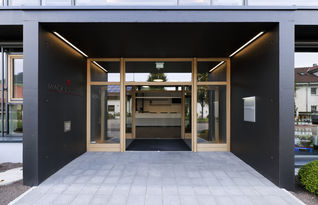 Foto: Die Außenhülle des Eingangsbereiches ist aus einem schwarzen Betonrahmen, der im Kontrast zu der hellen Holzeinfassung der Glasscheiben steht.