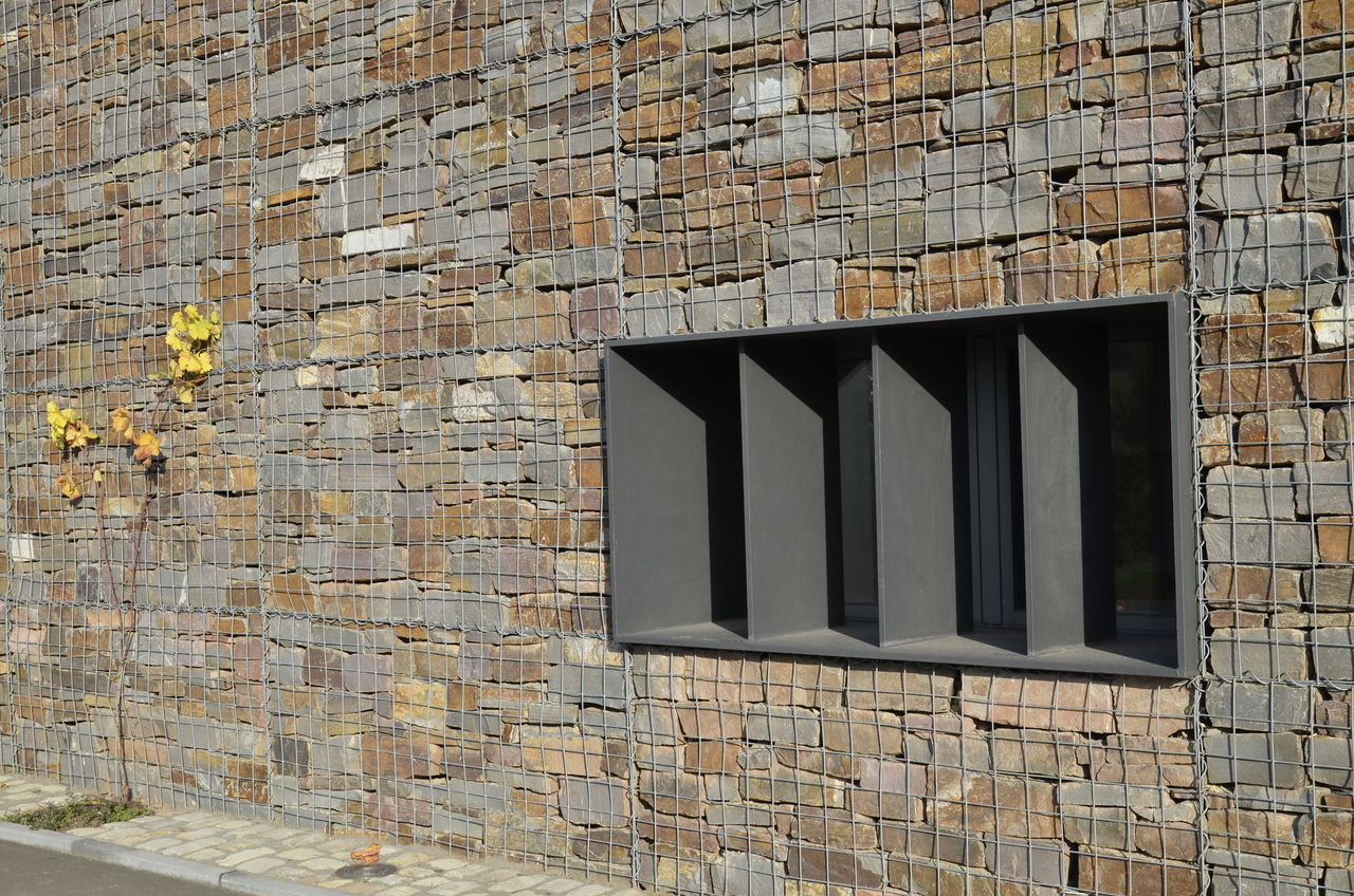 Fensteröffnung mit einem dunkelgrauen Massivrahmen durchbricht die Bruchsteinmauer des Wirtschaftsgebäudes.