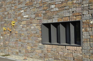 Fensteröffnung mit einem dunkelgrauen Massivrahmen durchbricht die Bruchsteinmauer des Wirtschaftsgebäudes.