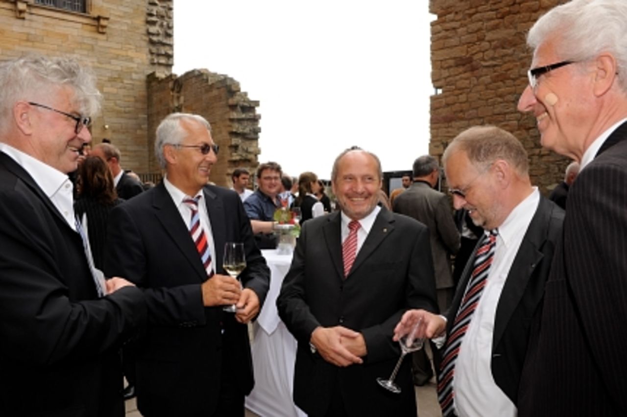Foto: Fünf Männer im Gespräch, im Hintergrund ein Loch in der historischen Mauer deutet die Ruine an.