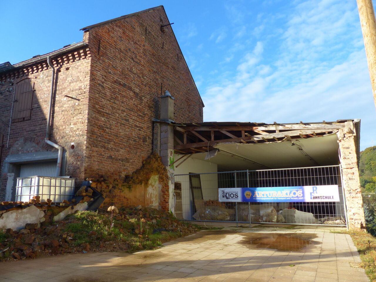 Links hohes fensterloses Gebäude (Stall) aus Ziegeln, daneben eine baufällige Garage mit Auffahrt, halb im Abbruch begriffen.