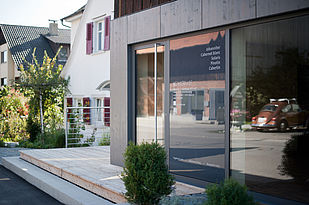 Foto: Blick auf den neuen Eingang mit großer Glastür, der in den neuen Teil der Holzfassade eingelassen ist.