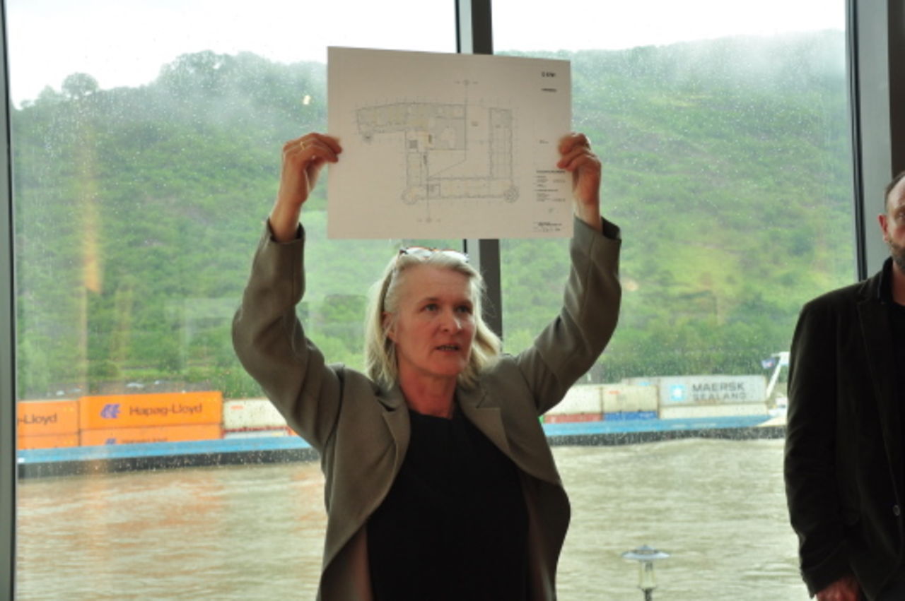 Architektin Constanze Altemüller präsentiert den Besuchern einen Plan, im Hintergrund fährt ein Frachtschiff auf dem Rhein vorbei