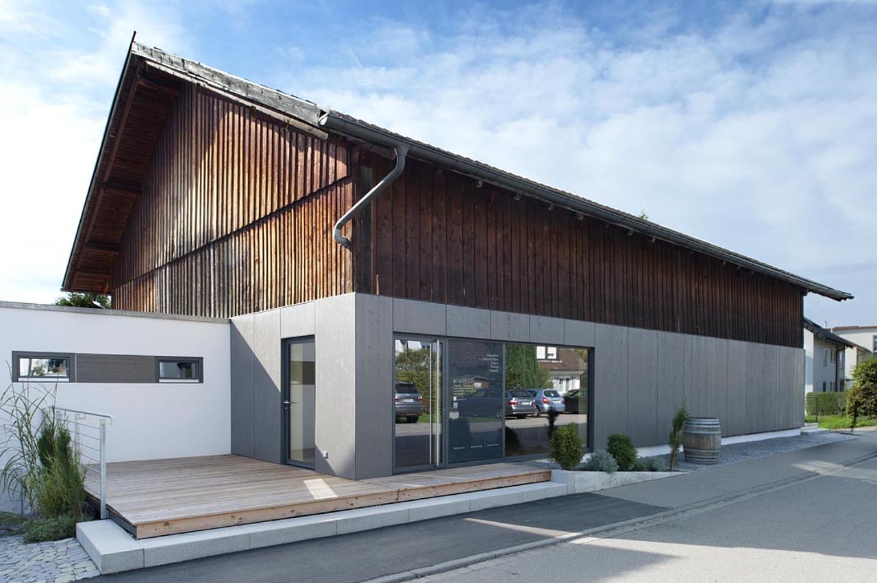 Foto: Alt trifft auf neu. Blick von der gegenüberliegenden Straßenseite auf die modernisierten Scheune die im Bereich des Erdgeschosses mit glatten, lichtgrauen Holzscheiben versehen wurde