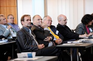 Foto: Blick von vorne in die erste Publikumsreihe: Sieben Menschen hören aufmerksam dem Redner außerhalb des Bildes zu.