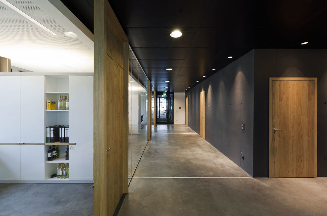 Foto: Blick auf den Gang, der zu den modernen Büroräumen führt. Die Wände sind aus dunkel eingefärbtem Sichtbeton, dies steht im Kontrast zu den rustikalen Türen aus hellem Holz.