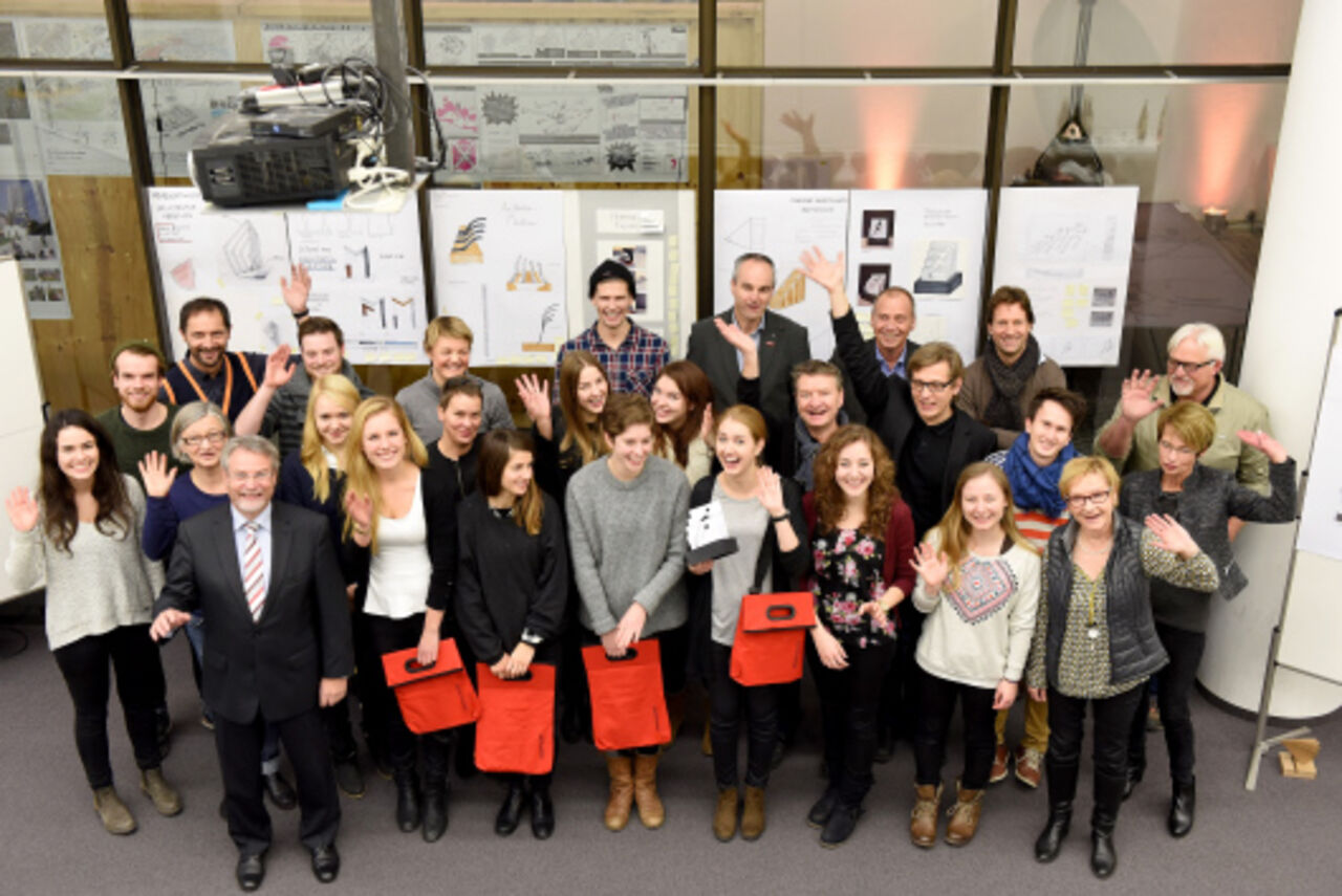 Die Gruppe aus Jurymitgliedern und Studenten lächelt und winkt in die Kamera, aufgenommen von schräg oben.