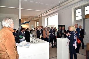 Der Blick in den ZUgangsbereich der Bauausstellung mit den großen Toren der ehemaligen Fahrzeughalle, am Pult steht Staatsministerin Doris Ahnen und spricht zu einer Gästegruppe von rund 50 Personen.