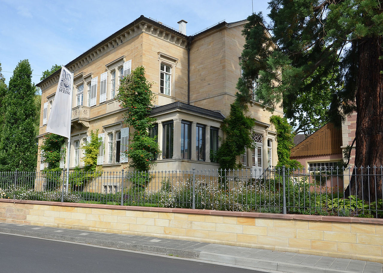 Foto: Blick auf eine Villa aus hellem Sandstein,die sich auf einem umzäunten, mit Bäumen bewachsenen Grundstück befindet.