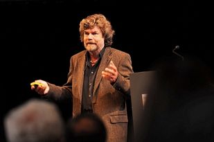 Foto: Messner vor dunklem Hintergrund auf der Bühne, vorne die schemenhaften Köpfe des Publikums