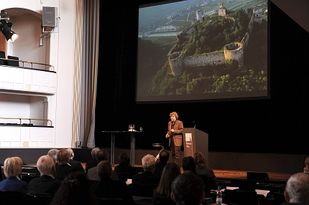 Foto: Der Saal in der Totalen. Messner vorne vor der Leinwand, das Publikum hört aufmerksam zu. Im Hintergrund die Leinwand mit einem der fünf Museen