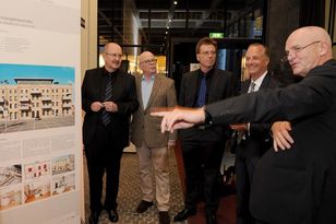 Fünf Herren stehen im Halbkreis vor einem Ausstellungsplakat, der Herr im Vordergrund zeigt auf das Plakat und erklärt etwas.