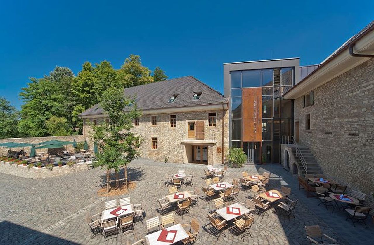 Foto: Blick in den Innenhof mit Außenbestuhlung. Der Boden ist passend zu der hellen Steinfassade mit hellem Stein gepflastert.