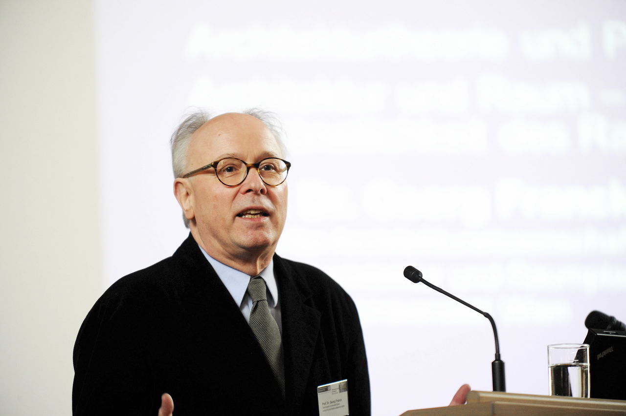 Älterer Herr mit Brille an einem Rednerpult mit Präsentation im Hintergrund.
