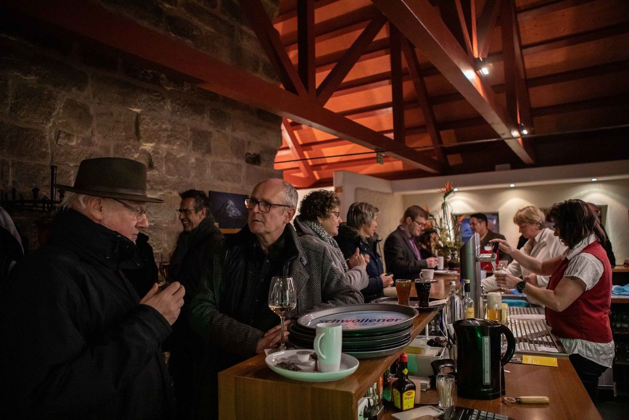 Blick in den Gastraum mit großer Theke und einigen Besuchern, die hier ein Getränk zu sich nehmen.