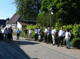 Dörfliche Straßensituation mit einer Gruppe von ca. 20 Personen, die sich im Sonnenschein Häuser ansehen.