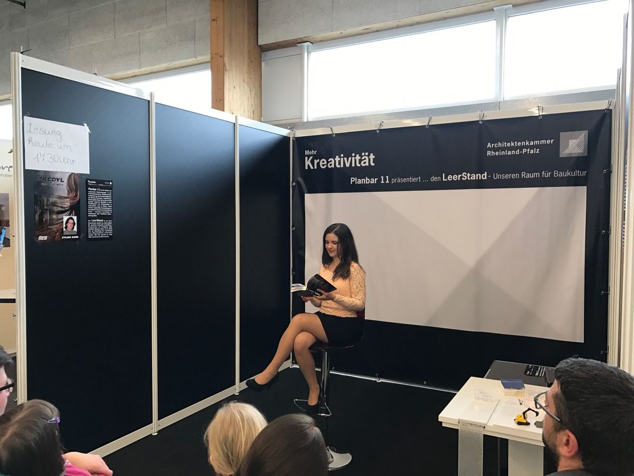 Der kleine Messestand, in dessen Mitte eine junge Frau auf einem Barhocker sitzt und aus einem Buch vorliest.