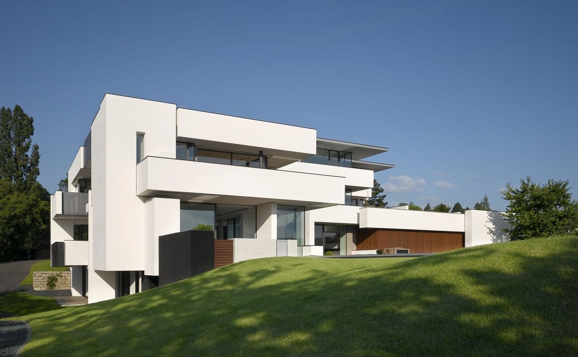 Blick auf eine weiße Villa auf einem Hang mit Wiese vor blauem Himmel.