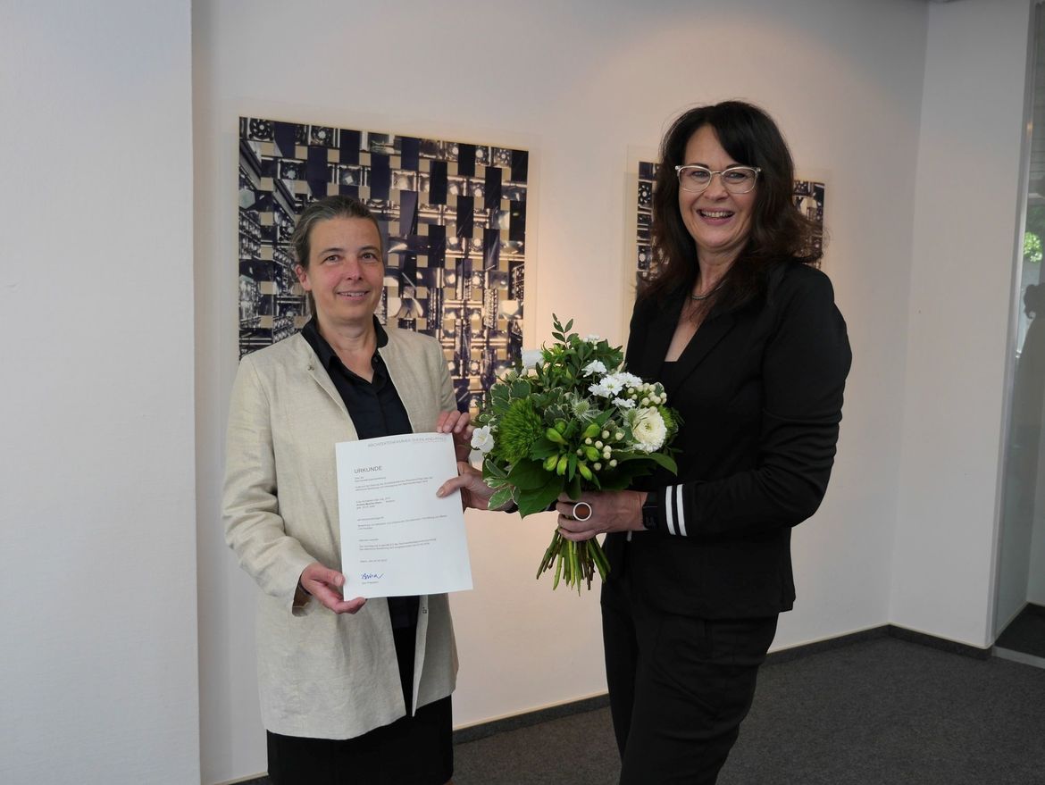 Vizepräsidentin Edda Kurz steht zusammen mit Andrea Mueller-Klein in der Landesgeschäftsstelle. Frau Kurz hält die Bestallungsurkunde in den Händen, Frau Mueller-Klein einen BlumenstraußRundstempel und Sachverständigenausweis.