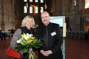 Staatsministerin Doris Ahnen mit einem Blumenstrauß vor dem bestuhlten Saal der Christuskirche, Sie und Gerold Reker blicken fröhlich in die Kamera.