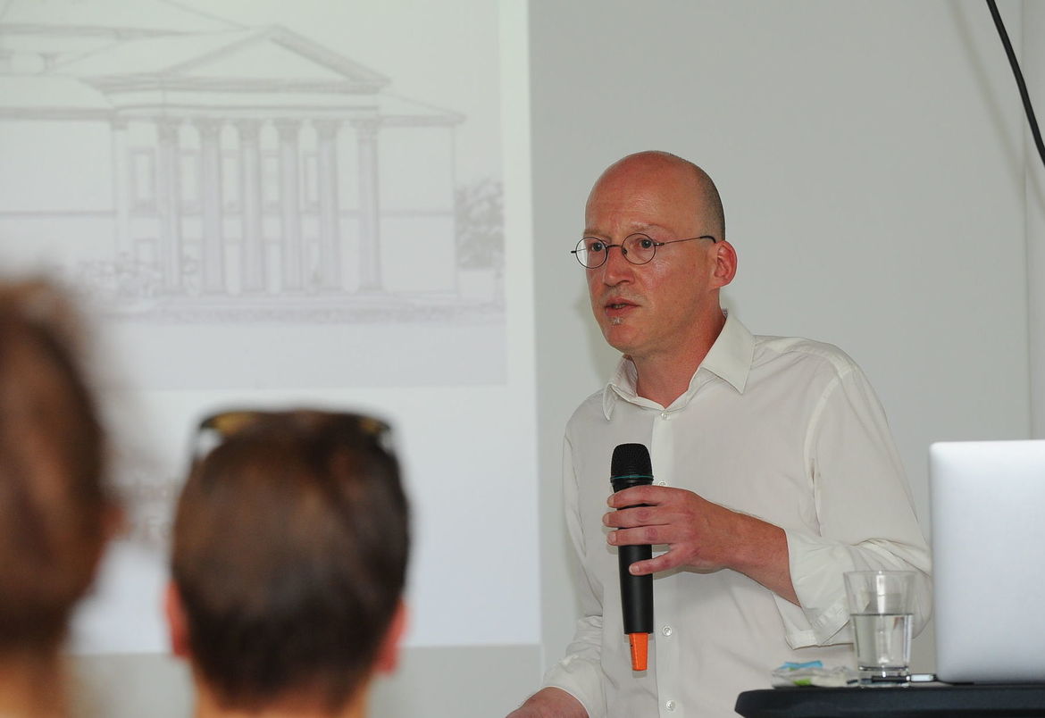 Portraitfoto von Theaterwissenschaftler Sascha Salzig, im Hintergund die Beamerprojektion.