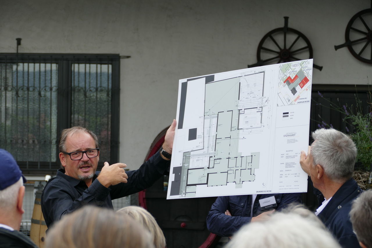 Architekt hält Plan hoch, auf dem er Besuchern Planungen erläutert