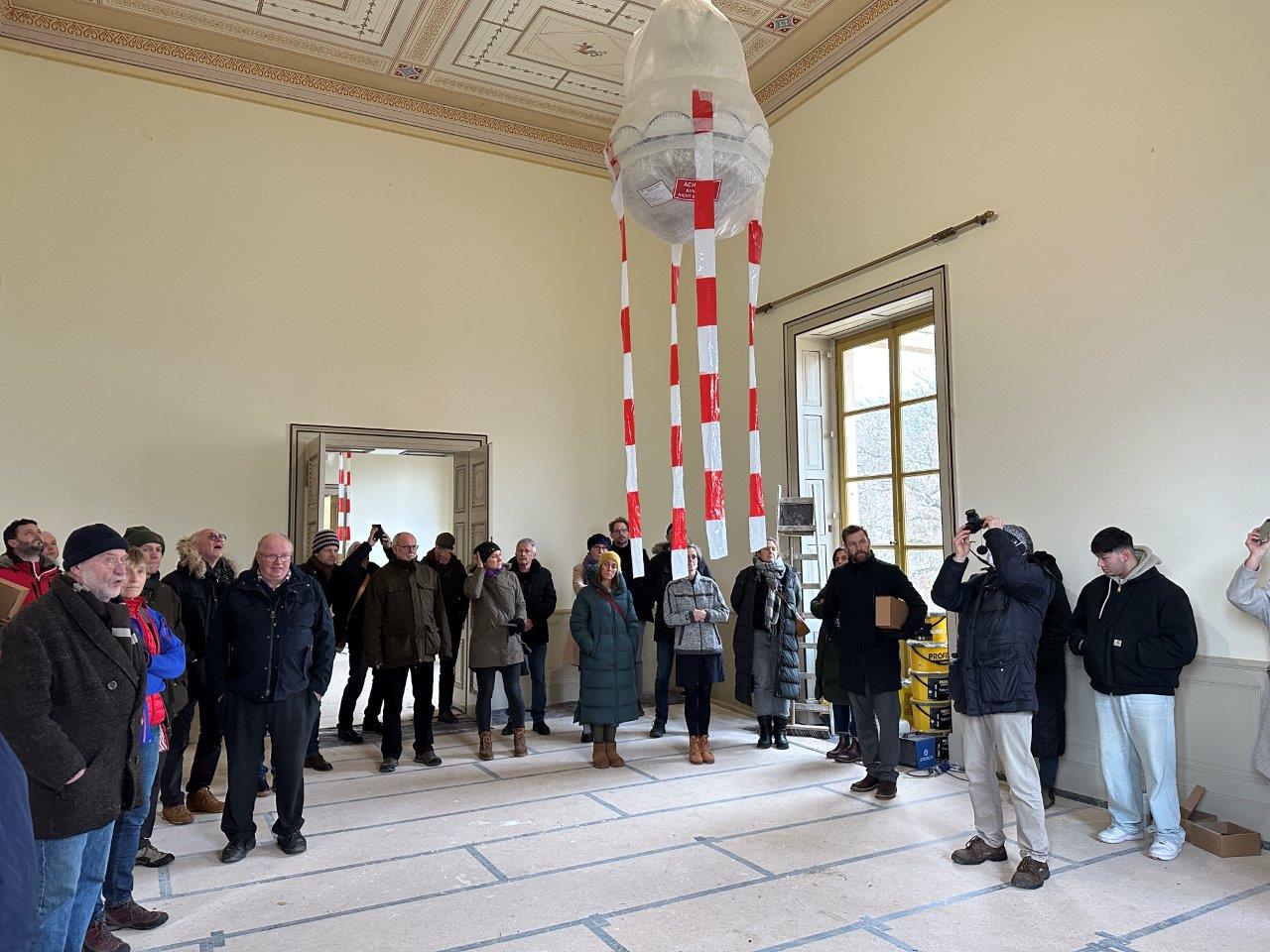 Eine große Gruppe im großen Saal, der Kronleuchter ist noch verhüllt, rot weiße Flatterbänder hängen herunter.