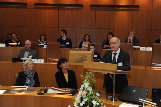 Zuhörer sitzen im runden Ratssaal aus Holz und hören dem Referenten am Rednerpult zu.