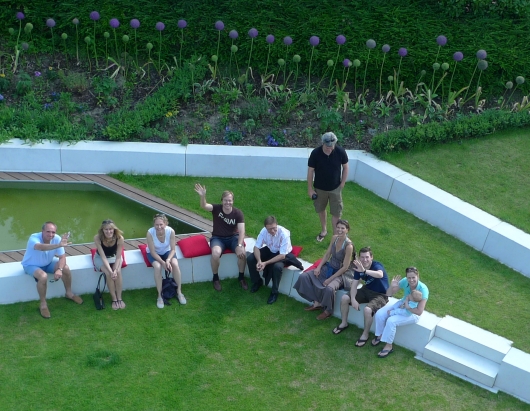 Foto: Annette Müller, Mainz Foto: Blick von oben in einen geometrisch gestalteten Garten mit Rasenflächen und schräg-winkligen Betonstufen. Auf den Stufen sitzen Menschen und winken.